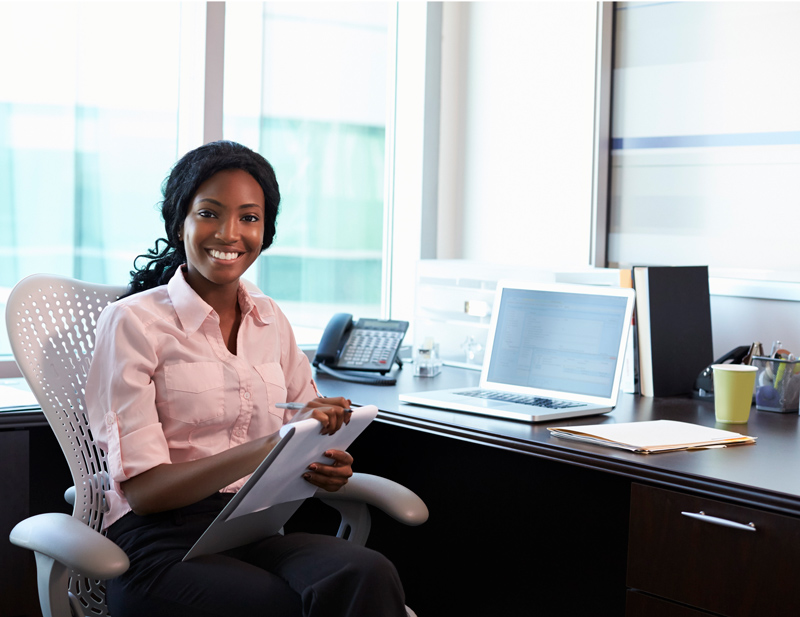 Woman sitting at a desk in an office, holding a clipboard and pen, smiling at the camera. A laptop, phone, and papers are on the desk.