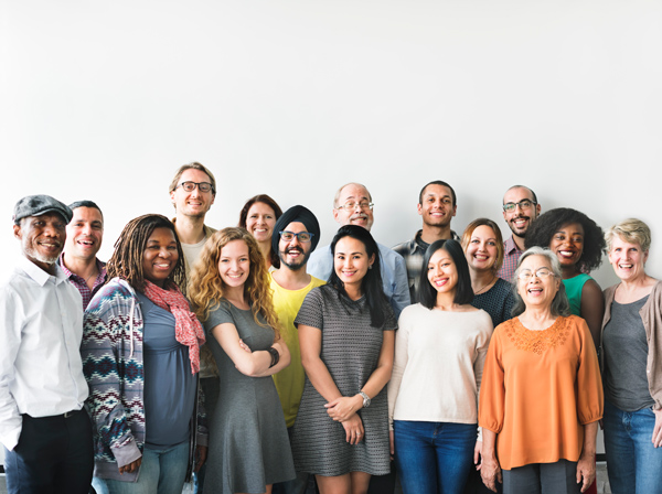 A group of sixteen diverse adults stand together in front of a plain white wall, smiling at the camera.