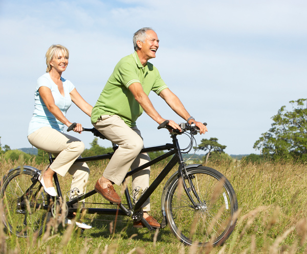 Two older adults ride a tandem bicycle through a grassy field on a sunny day. Both are smiling and dressed in casual light-colored clothing.