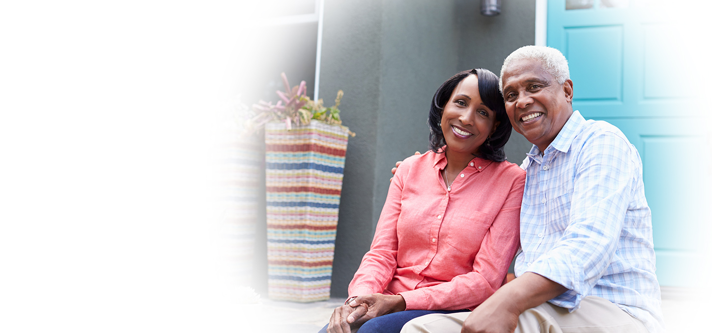 An older couple sits together and smiles on the front steps of their home, next to a colorful plant basket and a bright blue door.