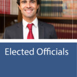 A man in a suit and red tie smiles at the camera, standing in front of bookshelves. The text below reads, “Elected Officials.”.