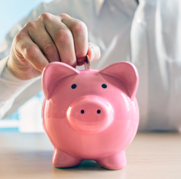 A person places a coin into a pink piggy bank sitting on a wooden surface.