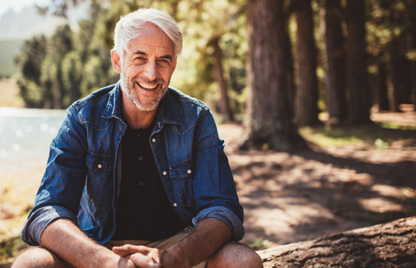 A man with gray hair and a beard sits outdoors on a log, smiling, with trees and sunlight in the background.