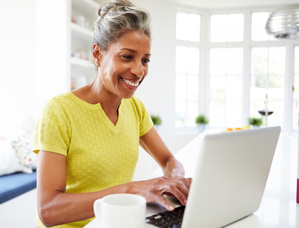 Smiling woman in a yellow shirt sits at a table and types on a laptop in a bright, modern kitchen.