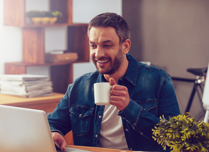 A man in a denim shirt sits at a desk, smiling at his laptop while holding a white mug. There are shelves and a potted plant in the background.