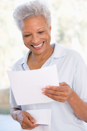 An older woman with short gray hair smiles while reading a piece of paper, holding an envelope in her other hand.