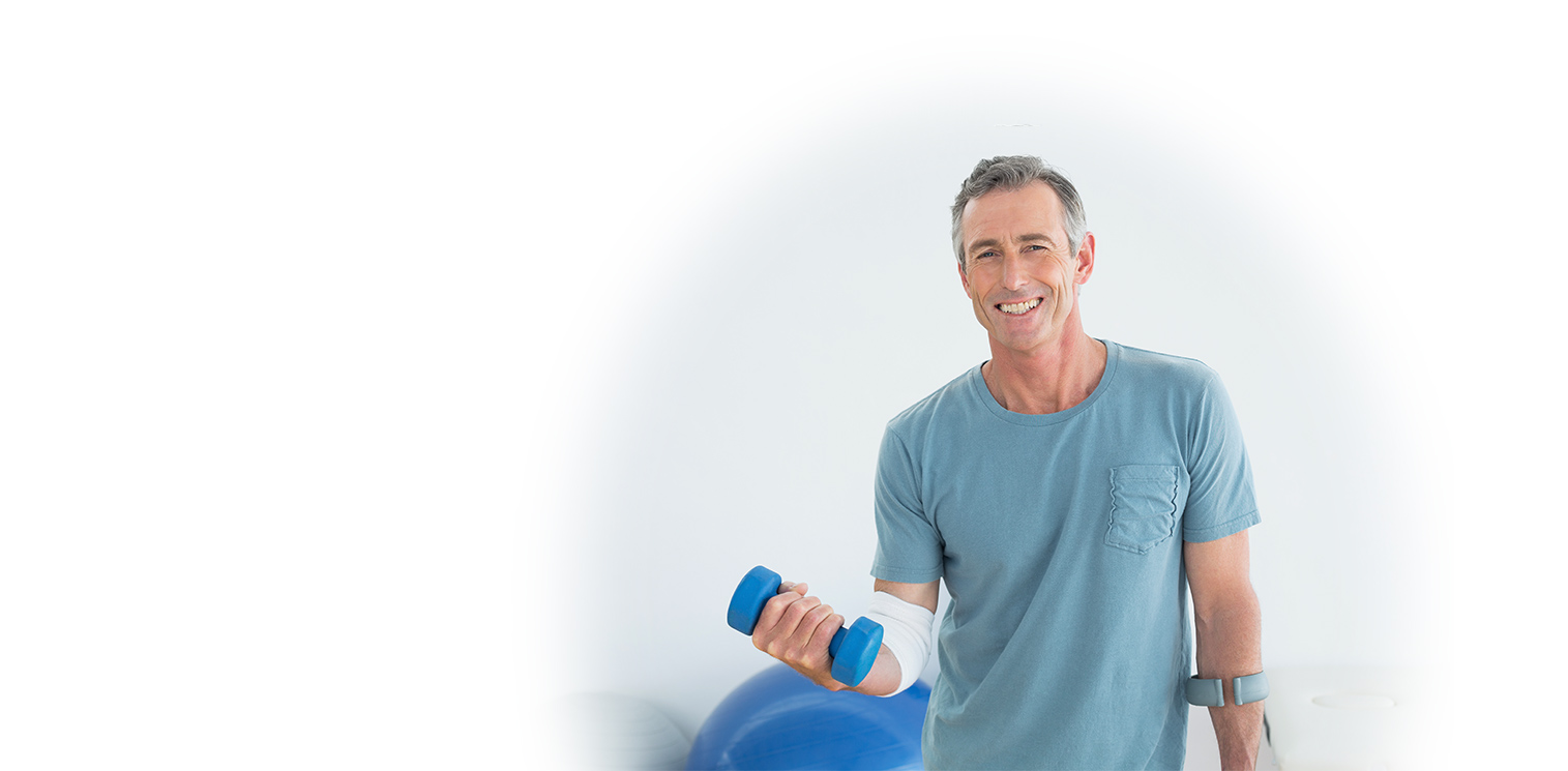 Man with a bandaged arm smiles while holding a blue dumbbell, standing indoors with exercise equipment in the background.