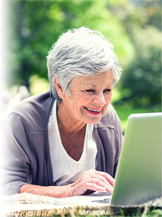 Older woman with short gray hair smiling while using a laptop outdoors, with green trees blurred in the background.