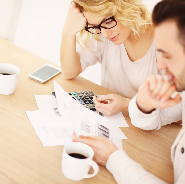 Two people sit at a table with documents, a calculator, coffee cups, and a smartphone, appearing focused while reviewing paperwork.