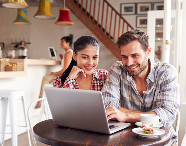 Two people sit at a café table, looking at a laptop and smiling. A coffee cup and dessert are on the table. A person is visible in the background near the counter.