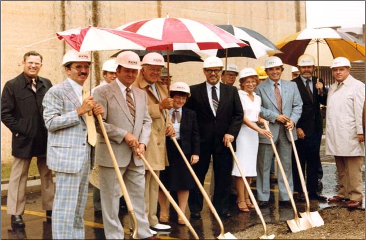 A group of people in formal attire and hard hats hold shovels at a groundbreaking ceremony, standing under umbrellas on a wet day.