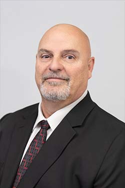 Bald man with a gray goatee wearing a black suit, white shirt, and patterned tie, posing for a professional headshot against a plain background.