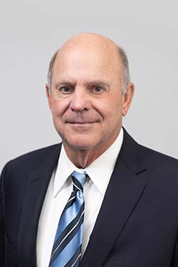 A middle-aged man wearing a dark suit, white shirt, and striped blue tie, posing against a plain light gray background.