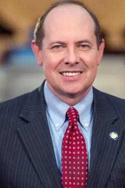A middle-aged man wearing a dark pinstripe suit, light blue shirt, and red patterned tie, smiling at the camera against a blurred background.