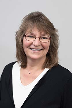 Smiling woman with shoulder-length brown hair, wearing glasses, a white top, and a black cardigan, posed in front of a plain light background.