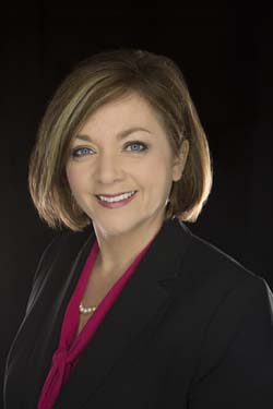 Woman with short light brown hair wearing a black blazer, magenta blouse, and pearl necklace, smiling in front of a dark background.