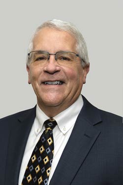 Older man with short white hair and glasses wearing a dark suit, white shirt, and patterned tie, smiling in front of a plain light gray background.