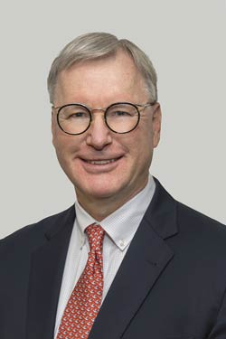 A man with short gray hair, wearing round glasses, a dark suit, white shirt, and red patterned tie, smiles at the camera against a plain light background.