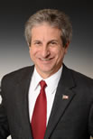 A man in a dark suit, white shirt, and red tie is smiling at the camera. He has gray hair and is wearing a small American flag pin on his lapel.