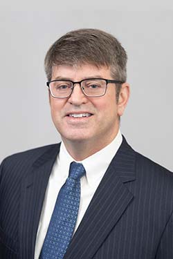 A man wearing glasses and a dark pinstripe suit with a white shirt and blue tie, posing in front of a plain light gray background.