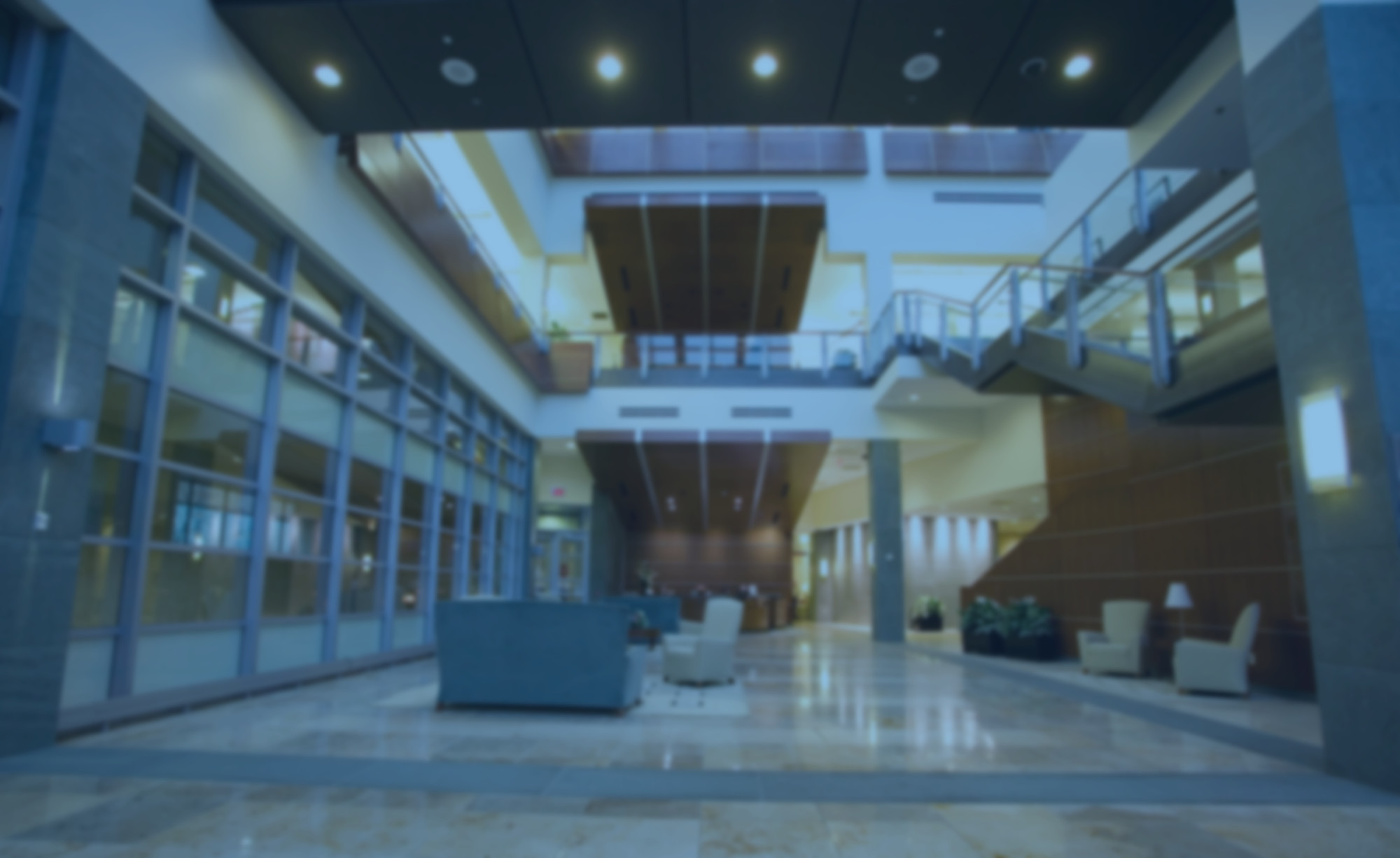 Blurred image of a modern office building lobby with seating areas, large windows, a staircase, and marble flooring.