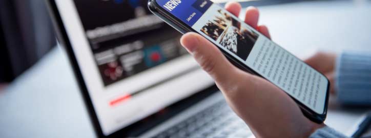 A person holds a smartphone displaying a news article, with an open laptop showing a blurred webpage in the background.
