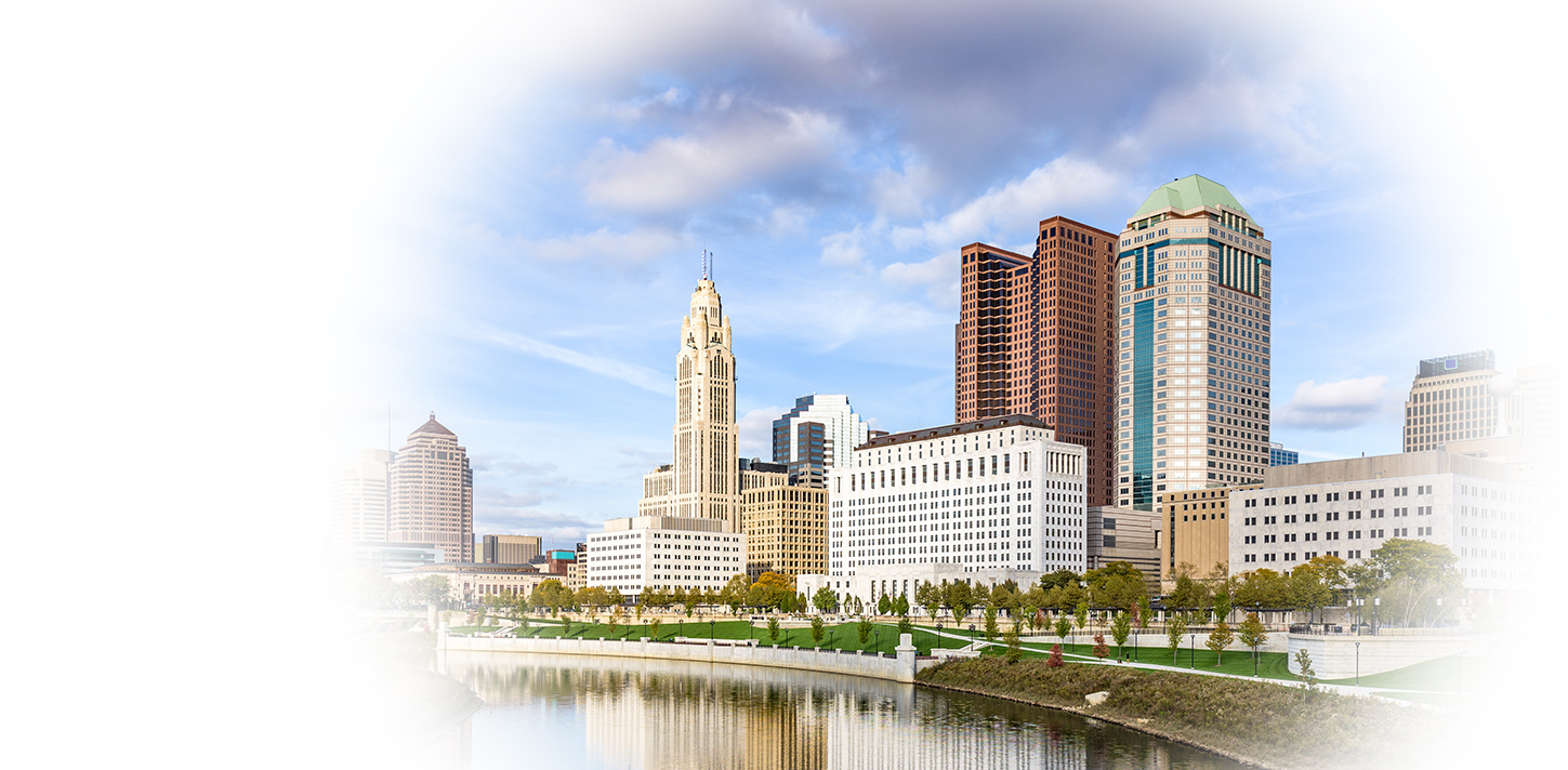 Downtown city skyline with tall office buildings, a river in the foreground, and a partly cloudy sky.