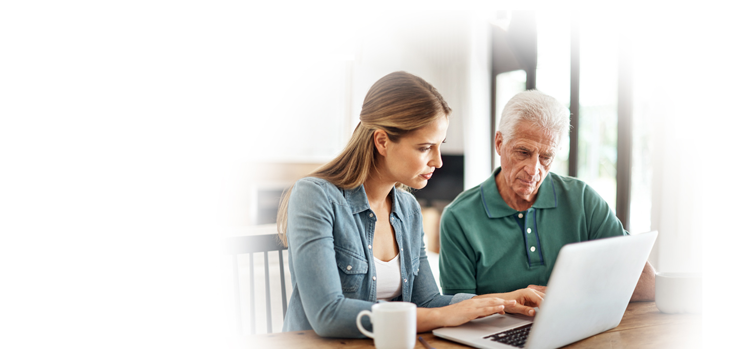 A young woman and an older man sit at a table looking at a laptop together, with coffee mugs nearby.