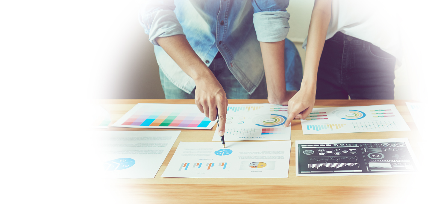 Two people stand at a table, reviewing and pointing at printed charts and graphs spread out on the surface.