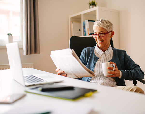 An older woman sits at a desk, reading a newspaper and holding a mug, with a laptop and notepad in front of her in a bright, modern office.
