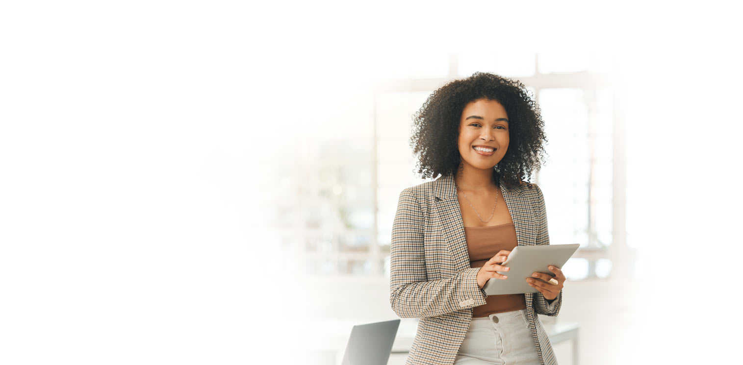 A woman stands indoors, smiling and holding a tablet. She wears a plaid blazer over a brown top and beige pants. The background is bright and softly blurred.
