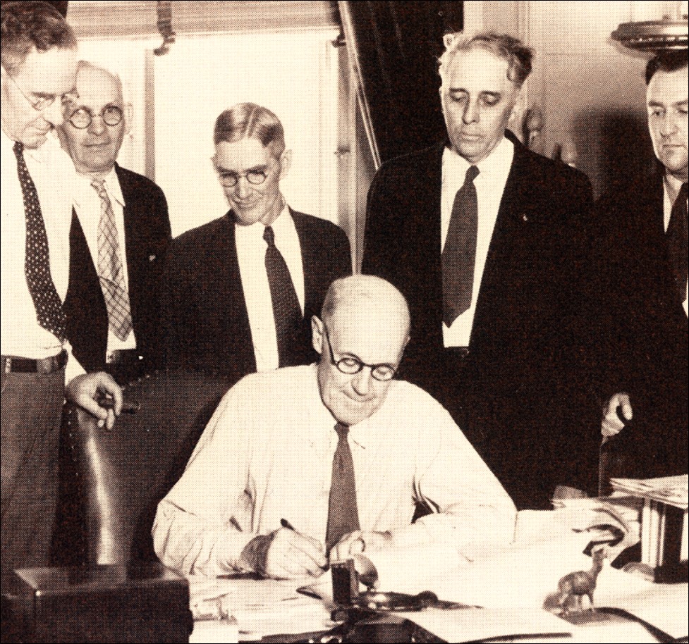 A group of men in suits observe as a seated man signs a document at a desk in an office setting.