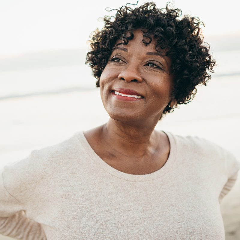 A woman with short curly hair smiles while standing outdoors, wearing a light-colored sweater. The background appears to be a beach or coastal area.