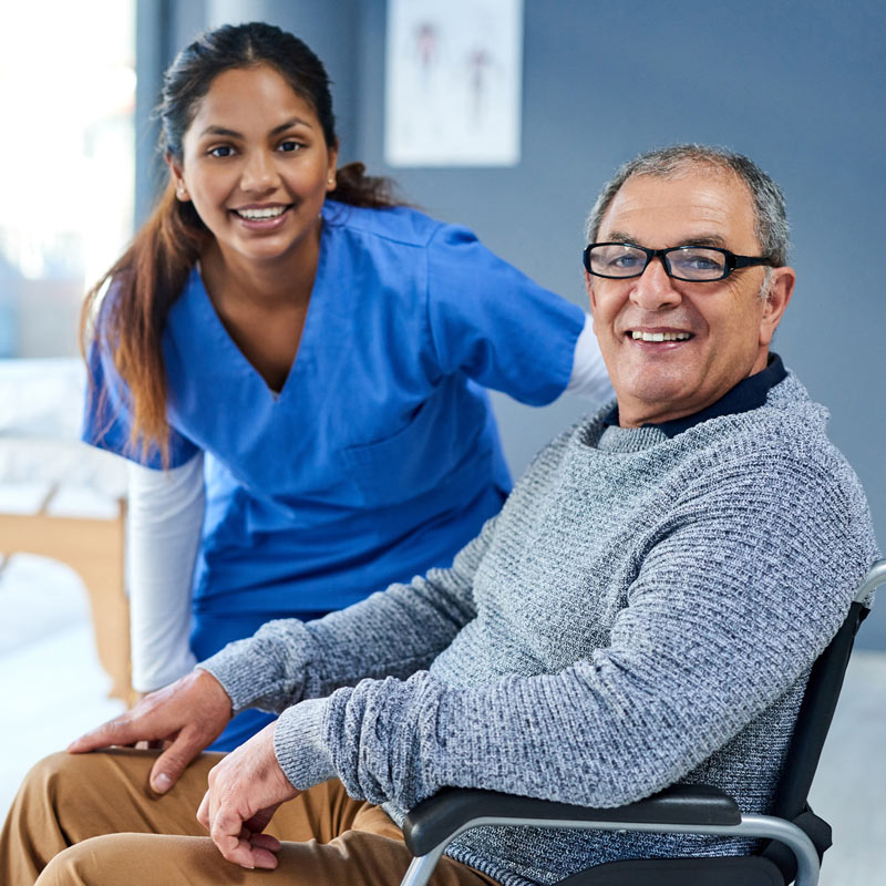 A nurse in blue scrubs stands next to an older man in a wheelchair, both smiling at the camera in a medical or care facility.