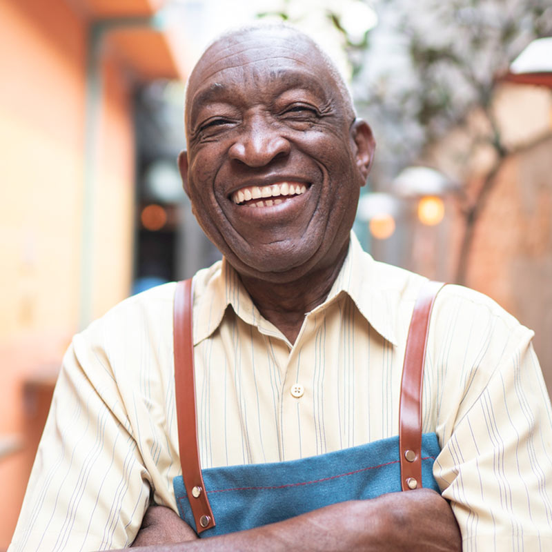 Older man wearing a striped shirt and apron stands outdoors with arms crossed, smiling broadly at the camera.