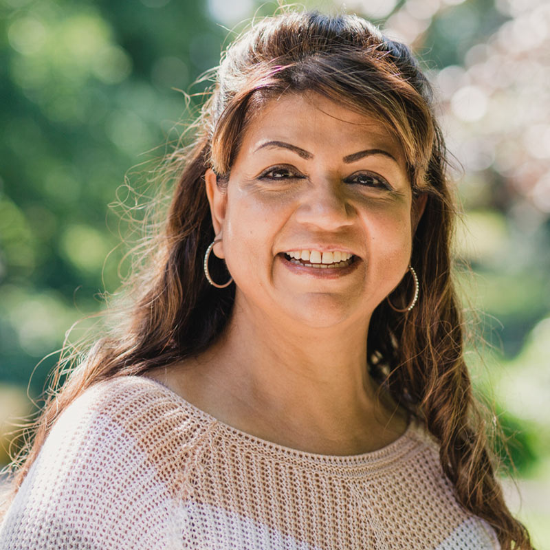 A woman with long dark hair and hoop earrings smiles outdoors, wearing a light knit top with a blurred green background.