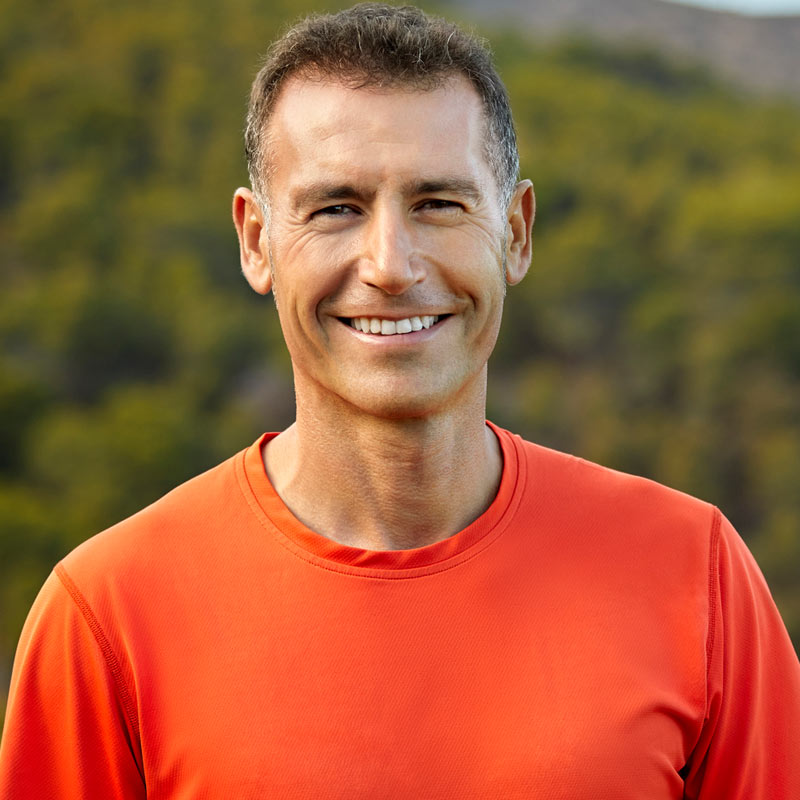 Man smiling outdoors wearing an orange shirt, with trees and hills blurred in the background.