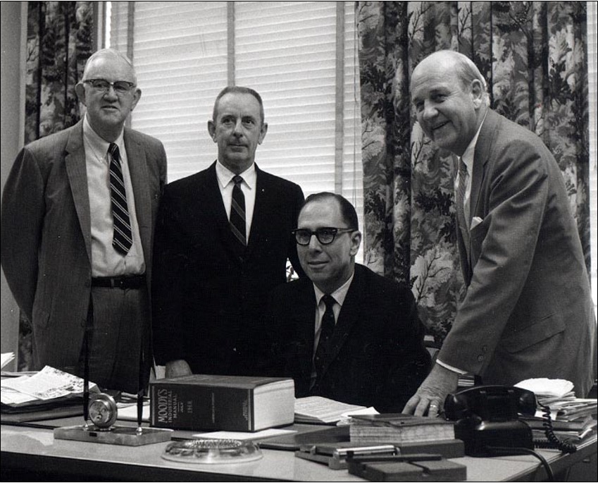 Four men in suits pose in an office setting; three stand behind a desk while one is seated. The desk is cluttered with papers, books, and office supplies.