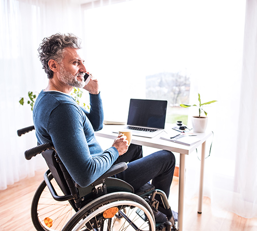 Man in a wheelchair talking on a smartphone while holding a mug, seated at a desk with a laptop, notepad, and potted plant in a bright room.