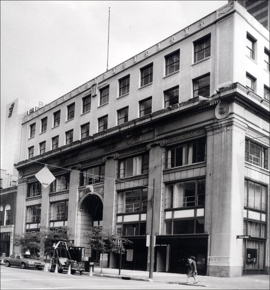 A four-story historic building with large arched entryway and classical architectural details stands on a city street, with pedestrians and parked cars in front.