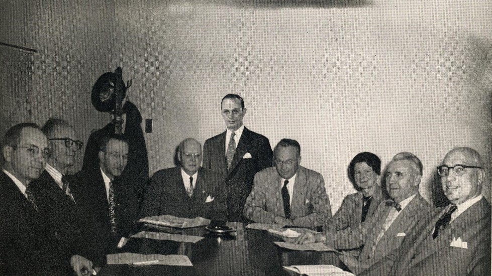 Nine people in suits sit and stand around a table in a meeting room, with documents in front of them and a coat rack in the background.