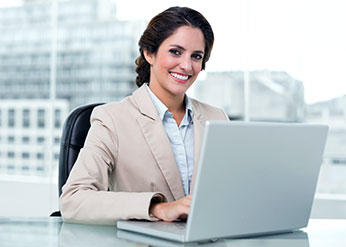 Woman in business attire sitting at a desk, smiling at the camera, and using a laptop in a modern office setting.