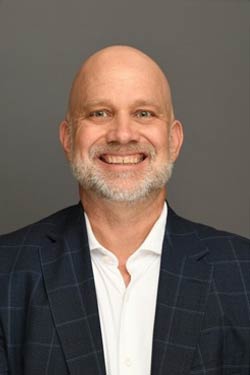 A middle-aged man with a bald head and gray beard, wearing a dark plaid suit jacket and white shirt, smiling in front of a plain gray background.