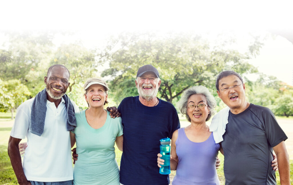 Five older adults stand outdoors in casual athletic wear, smiling at the camera in a park setting with trees in the background.