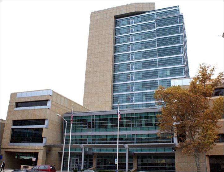 A modern multi-story office building with glass windows and beige exterior, two flagpoles in front, and a tree on the right side.