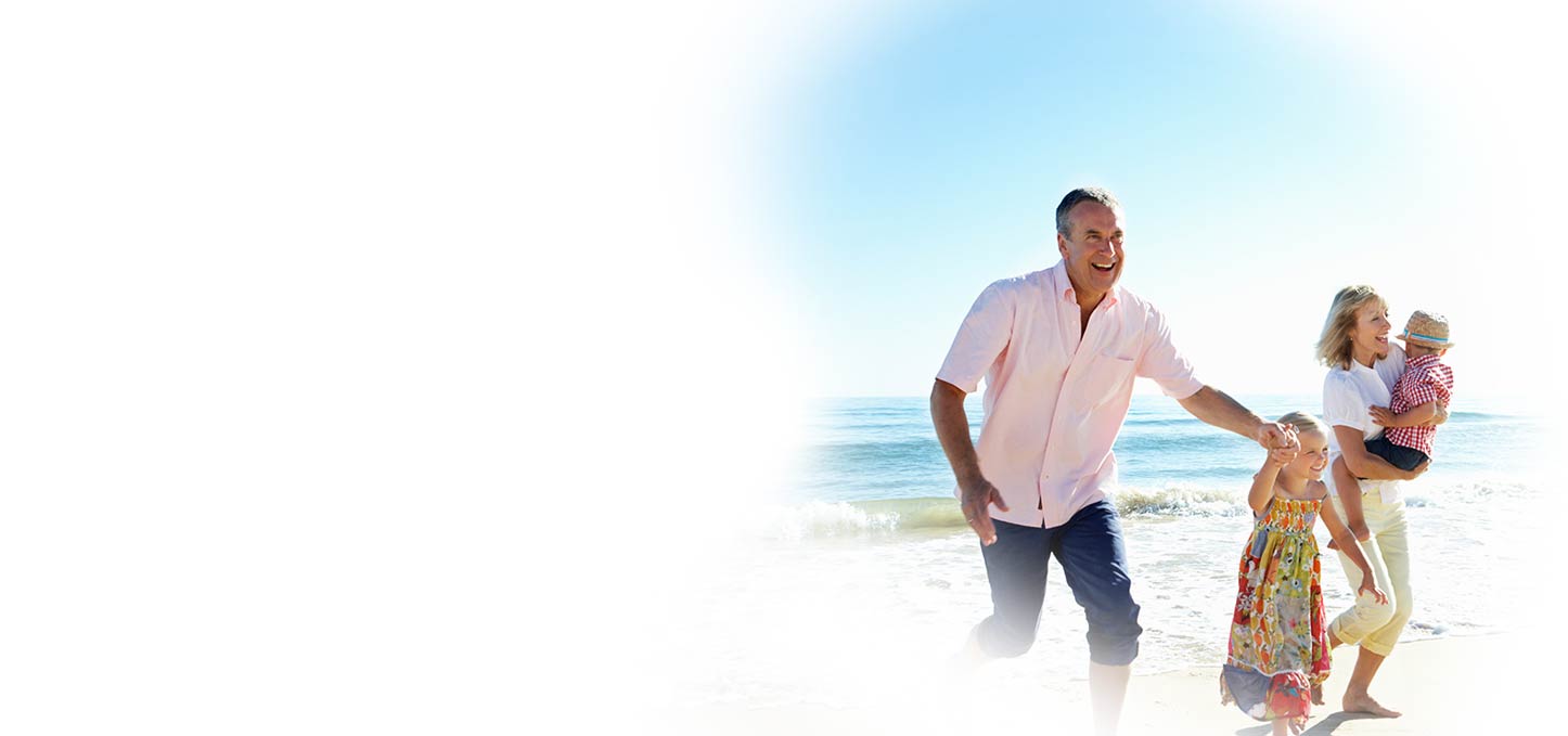 A man, woman, and two young children walk and play together at the beach on a sunny day, with ocean waves in the background.